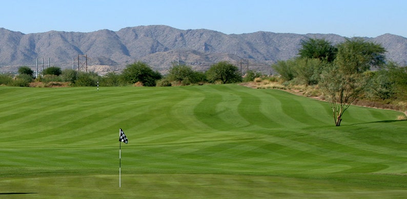 May include: A golf course with a green putting green and a flag in the distance. The green is surrounded by a grassy area and a mountain range in the background.