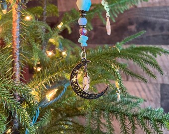 Op de afbeelding: Een halve maanvormige ornament met een kristallen hanger. De ornament is gemaakt van metaal en heeft een kralenketting met blauwe, paarse en heldere kralen.