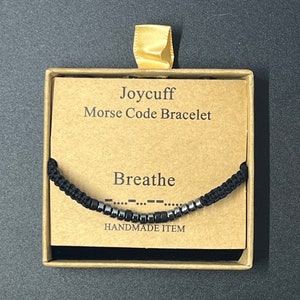 May include: A black string bracelet with silver beads spelling out "Breathe" in Morse code. The bracelet is in a small box with a brown paper label that reads "Joycuff Morse Code Bracelet" and "Handmade Item".