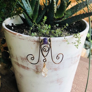 May include: A wire plant hanger with a purple amethyst bead and a clear quartz crystal hanging from it. The hanger is attached to a white ceramic pot with a succulent plant growing in it.