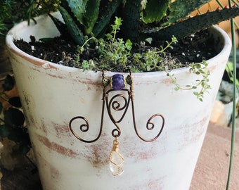 Op de afbeelding: Een plantenhanger van draad met een paarse amethistkraal en een helder kwartskristal dat eraan hangt. De hanger is bevestigd aan een witte keramische pot met een vetplant die erin groeit.
