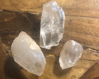 May include: Three clear quartz crystals of varying sizes on a wooden surface.
