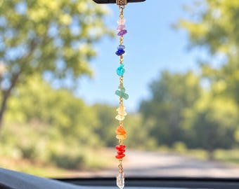 Puede incluir: Un adorno colgante para el espejo retrovisor del coche con una cadena dorada vertical que presenta siete piedras preciosas pulidas de colores en secuencia de arcoíris, rematada con una cuenta metálica y un colgante de cristal.