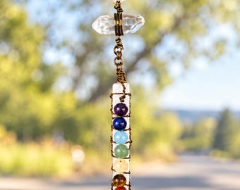 Puede incluir: Un adorno colgante para coche con una punta de cuarzo transparente en la parte superior, unida por una cadena de metal color bronce a una varita de selenita blanca envuelta en alambre. Siete pequeñas cuentas esféricas de colores están alineadas.