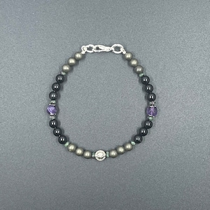 Puede incluir: Un brazalete con cuentas negras, verdes y moradas, un cierre plateado y un dije plateado.