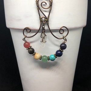 May include: A white ceramic planter with a wire hanger and a crystal point charm. The hanger is decorated with a rainbow of seven chakra beads.