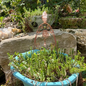 Puede incluir: Un soporte de cristal de alambre de cobre con un cristal rosa en la parte superior y cristales verdes en el alambre. El soporte está en una maceta azul con plantas verdes.
