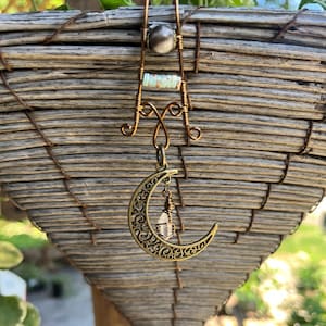 May include: A decorative hair accessory crafted from copper-toned wire featuring a central silver-colored bead and a row of small, pale green cylindrical beads. A filigree crescent moon charm hangs from the wire frame with a small clear crystal.