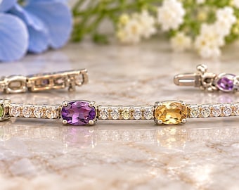 Può includere: Un bracciale in metallo color argento con una serie di pietre preziose ovali multicolori, tra cui pietre blu, verdi, viola, gialle e rosse. Il bracciale è impreziosito da piccole pietre rotonde trasparenti incastonate lungo tutta la fascia.