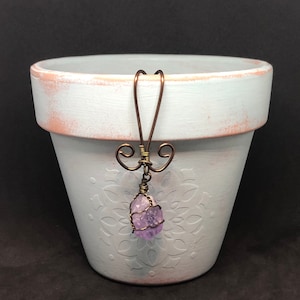 May include: A light blue ceramic planter with a white embossed design and a purple amethyst crystal hanging from a copper wire.