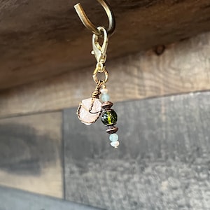 Pode incluir: Um berloque dourado com fecho lagosta, apresentando um cristal de quartzo rosa enrolado em arame e um grupo de contas verdes e azuis. O berloque está suspenso de um gancho curvo, mostrando um design de joia artesanal.