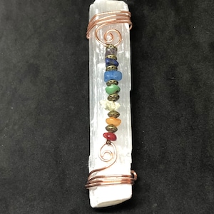 May include: A white selenite wand with copper wire wrapped around it. The wand has seven chakra beads strung on the wire, each bead representing a different chakra color.