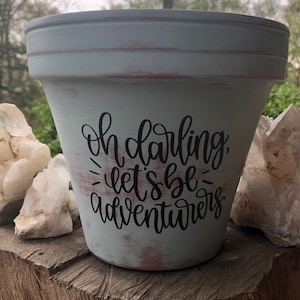 May include: A light blue terracotta planter with a distressed finish. The planter has black lettering that reads "Oh darling, let's be adventurers".