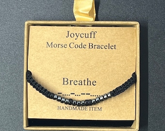 May include: A black string bracelet with silver beads spelling out "Breathe" in Morse code. The bracelet is in a small box with a brown paper label that reads "Joycuff Morse Code Bracelet" and "Handmade Item".