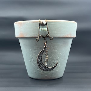 May include: A small, light blue terracotta pot with a decorative, wire-wrapped crescent moon charm hanging from the rim. The charm features a clear crystal and intricate metalwork.