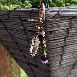 May include: A handmade crystal charm with a gold-colored clasp. The charm features a clear quartz crystal, green and black beads, and copper wire accents. The charm is hanging in front of a gray woven basket with green plants.