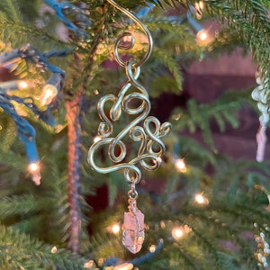 Op de afbeelding: Gouden draadversiering in de vorm van het getal 2023 met een roze kristal dat eronder hangt. De versiering hangt aan een kerstboom.