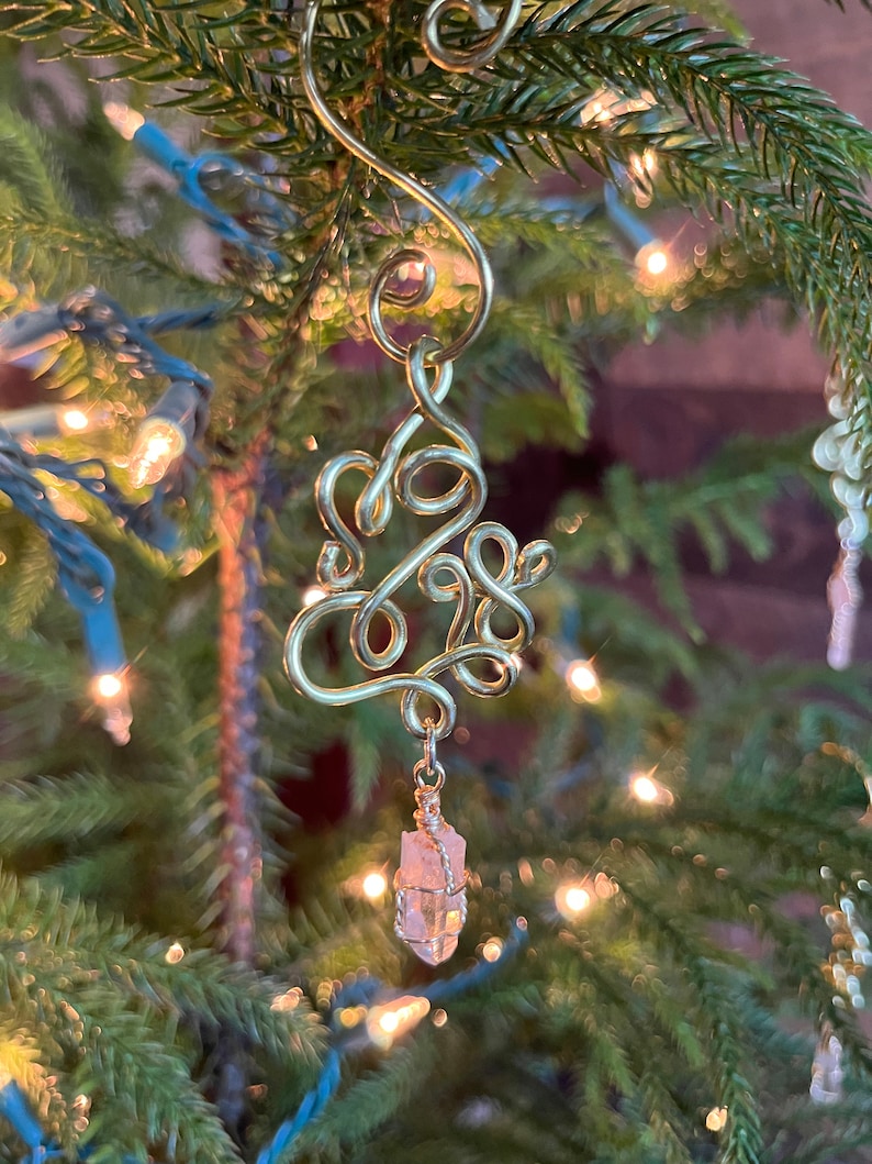 Op de afbeelding: Gouden draadversiering in de vorm van het getal 2023 met een roze kristal dat eronder hangt. De versiering hangt aan een kerstboom.