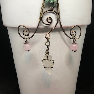 May include: A wire plant hanger with a decorative swirl design. The hanger has three hanging points, each with a pink crystal bead. One hanging point has a clear quartz crystal.