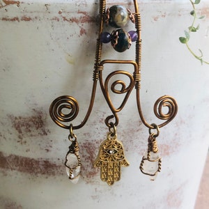 以下が含まれることがあります： 青い目をつけた金のハmsaの手のチャームが、透明な水晶と緑と紫のビーズが付いたワイヤーフレームからぶら下がっています。