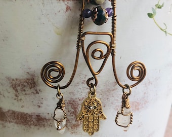Op de afbeelding: Een gouden hamsa-handhanger met een blauw oog, hangend aan een draadframe met heldere kwartskristallen en groene en paarse kralen.