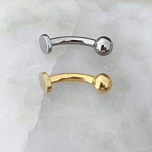 May include: Two curved eyebrow rings. One is silver-colored, and the other is gold-colored. Each ring has a flat disc on one end and a small ball on the other. The jewelry is displayed on a white, textured surface.