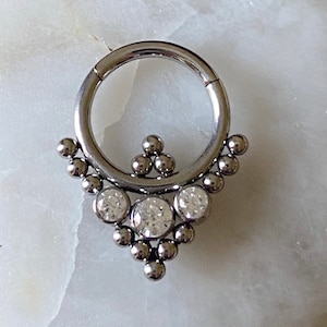 May include: A silver body jewelry piece with a circular hoop and a dangling, triangular design featuring three clear gemstones.