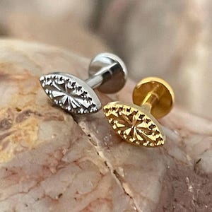 May include: Two stud earrings with an oval, starburst design. One earring is silver-coloured, and the other is gold-coloured. The earrings are displayed on a rough, brown and tan stone surface.