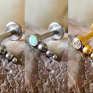 May include: Three different styles of body jewellery with a small gem and a row of beads. The first is silver with a clear gem, the second is silver with a white opal gem, and the third is gold with a clear gem.