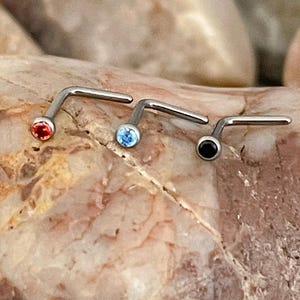 May include: Three nose studs with L-shaped posts. Each stud has a different coloured gemstone: red, light blue, and black. The studs are made of silver-coloured metal and are displayed on a textured rock surface.