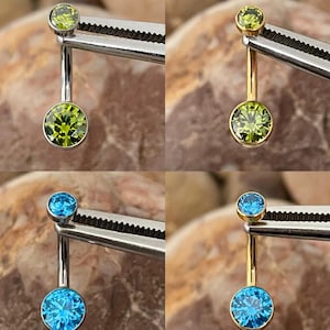 May include: Four belly button rings. Two have a silver-colored bar with a small and large green gemstone. The other two have a gold-colored bar with a small and large blue gemstone. The jewelry is held by a silver-colored tool.