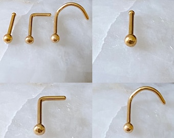 May include: Five gold nose rings with a ball end. The rings are in different styles, including straight, L-shaped, and curved.