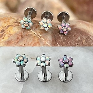 May include: Three surgical steel flower-shaped body jewellery pieces with opal-like stones. The flowers are in white, light blue, and pink.