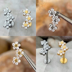 May include: Four different styles of body jewelry featuring a cluster of three flower-shaped charms with clear rhinestones. The charms are attached to a curved barbell post.