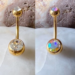 May include: Two gold belly button rings with clear and iridescent stones. The rings are on a white background.