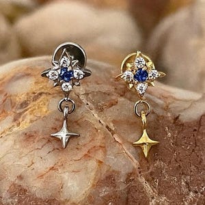 Peut inclure: Deux boucles d'oreilles en forme d'étoile, une argentée et une dorée, sont présentées. Chaque boucle d'oreille présente un motif en forme d'éclat d'étoile avec des pierres précieuses bleues et claires. Une petite étoile pend sous chaque boucle d'oreille, créant un thème céleste.