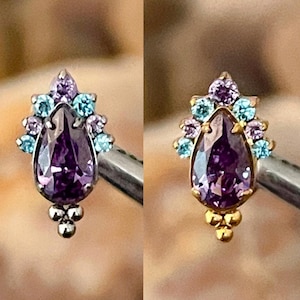 May include: Two ornate earrings, one silver and one gold, each featuring a large, teardrop-shaped purple gemstone. The earrings are adorned with smaller blue and purple stones, and a cluster of small spheres at the base.