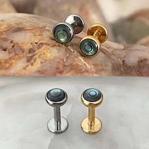 May include: Four abalone shell stud earrings are shown. Two are silver, and two are gold. Each earring has a flat, circular design with an iridescent abalone shell centre. The earrings are displayed on a light-coloured surface.