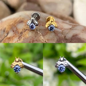 May include: Two small, decorative bee-shaped earrings. One is silver-toned, the other gold-toned. Each has a blue gemstone at the center. The earrings are shown on a natural stone surface and against a blurred green background.