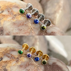 May include: Stud earrings with coloured gemstones. The image shows two rows of earrings, the top row with silver posts and the bottom row with gold posts. Each earring has a small, round gemstone in green, purple, blue, and yellow.