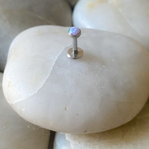 May include: A silver barbell with a small, round, iridescent opal gemstone. The barbell is on a white, smooth surface.