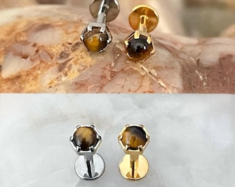 May include: Two pairs of stud earrings. One pair has a silver finish, and the other has a gold finish. Each earring features a round, tiger's eye gemstone set in a six-prong setting. The earrings are displayed on a textured surface.