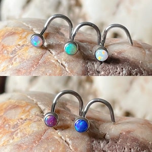 May include: Three pairs of nose rings with opal stones. The rings are made of surgical steel and have a curved shape. The opal stones are in various colours, including blue, purple, and white.