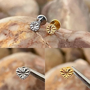 May include: Two small, decorative earrings. One is silver-colored, the other is gold-colored. Both have a flower-like design on the front. The earrings are shown on a rock surface. Two additional earrings are shown, one silver heart-shaped and one gold heart-shaped.
