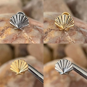 May include: Four seashell-shaped stud earrings in silver and gold tones. The earrings are displayed on a textured, light brown surface. The earrings have a detailed, fan-like design.