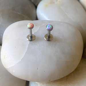 May include: Two silver body jewelry pieces with opal tops. The opal on the left is pink and the opal on the right is purple.