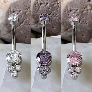 May include: Three belly button rings with different colored gemstones. The rings have a curved barbell design with a large round gem at the bottom and a smaller gem at the top. The gemstones are clear, purple, and pink.