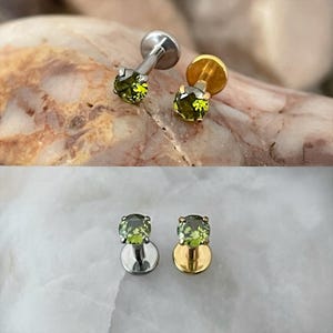 May include: Two small, stud earrings with a round, olive-green gemstone. One earring has a silver post and backing, while the other has a gold post and backing. The earrings are displayed on a textured, light-colored surface.