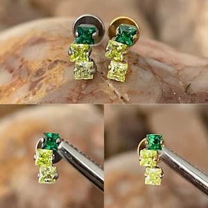 May include: A pair of stud earrings featuring a cluster of three square-cut gemstones in shades of green and yellow. The earrings have a silver and gold finish, with the gemstones arranged in a cascading design. The earrings are displayed on a textured surface.