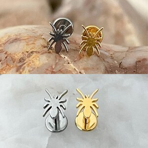 May include: A pair of spider-shaped stud earrings. One earring is silver-toned, the other is gold-toned. The earrings are displayed on a textured, light brown surface.